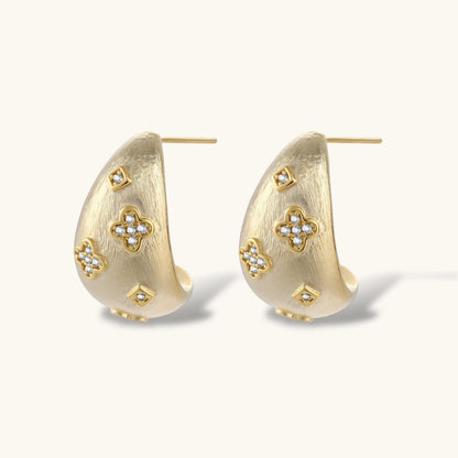 Curved gold-plated hoop earrings with four-leaf clover and diamond-shaped crystal details on white background